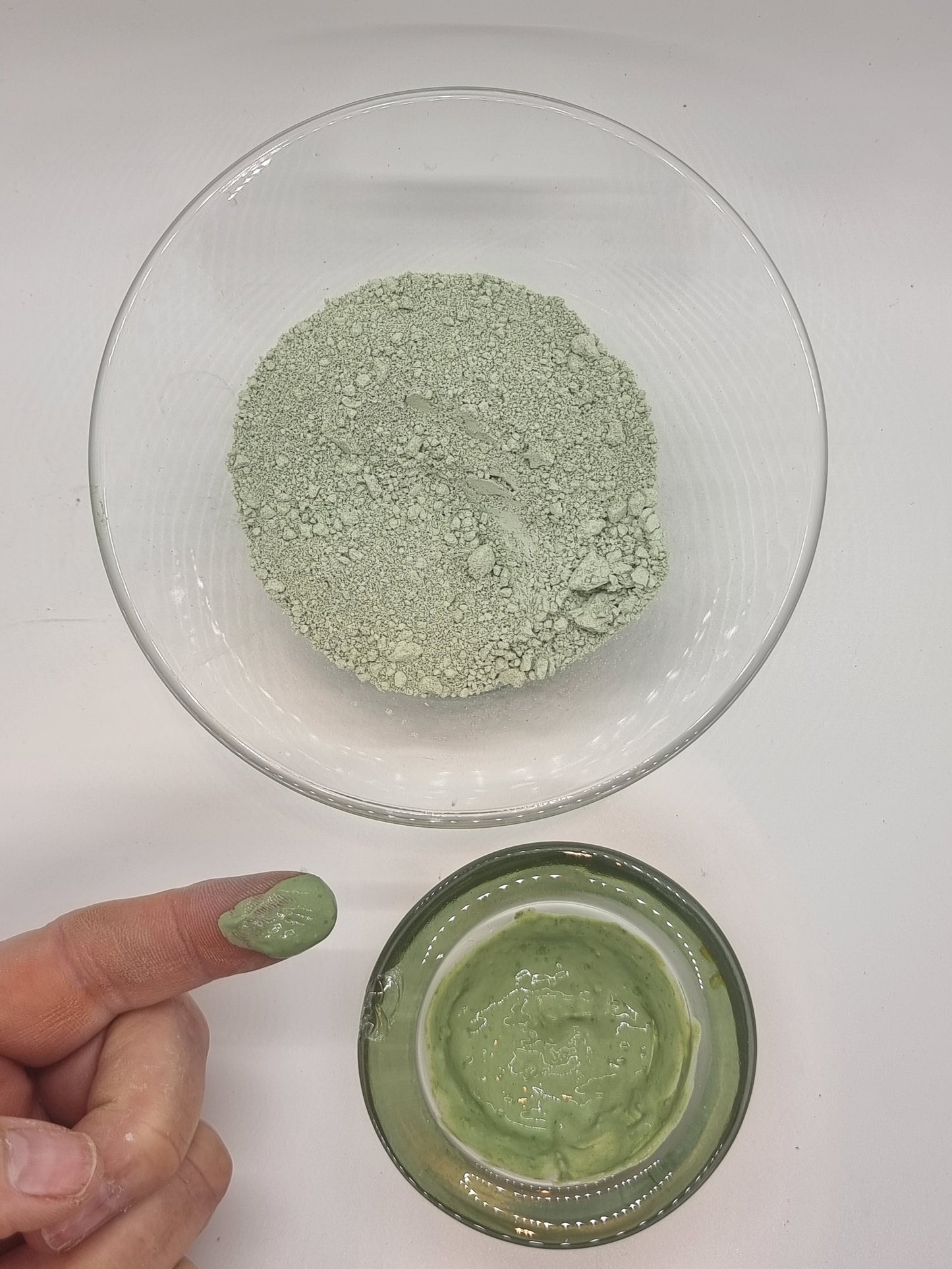 two glass bowls one containing dry fine french green clay that is a mid green colour, the other containing wet green clay that has a shiny appearance.  A hand and finger can be seen. The finger has a blob of shiny wet clay on it that is also green in colour.